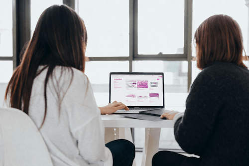 Twee vrouwen kijken naar het beeldscherm van een laptop en bespreken een contentstrategie in een lichte ruimte met veel natuurlijk zonlicht
