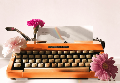Een oranje typemachine met daarin een beschreven bladzijde en een regenboogkleurige lichtreflectie en omringd door roze bloemen